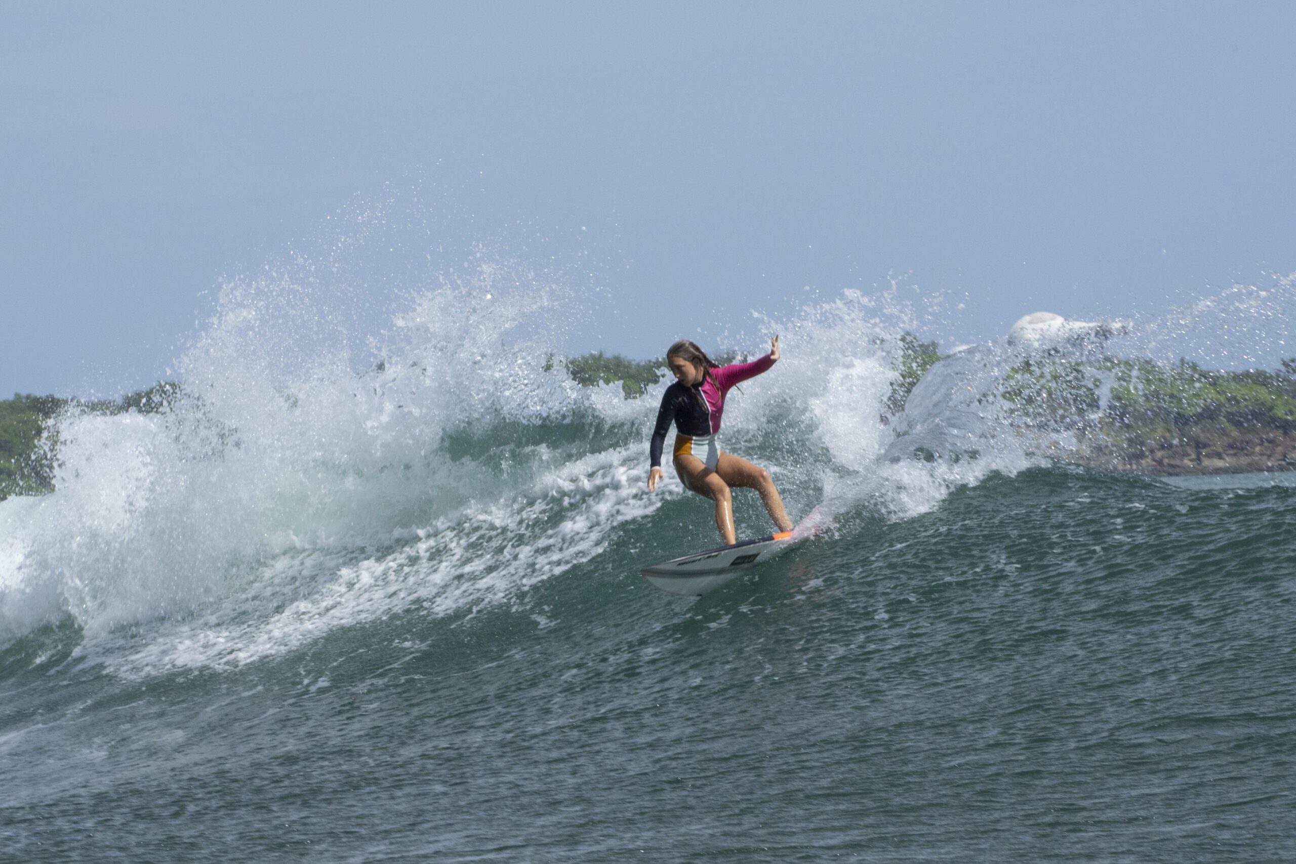 http://A%20surfer%20wearing%20a%20pink%20and%20black%20long-sleeve%20top%20and%20orange%20shorts%20rides%20a%20breaking%20wave,%20creating%20a%20spray%20of%20water,%20with%20greenery%20and%20a%20cloudy%20sky%20in%20the%20background.