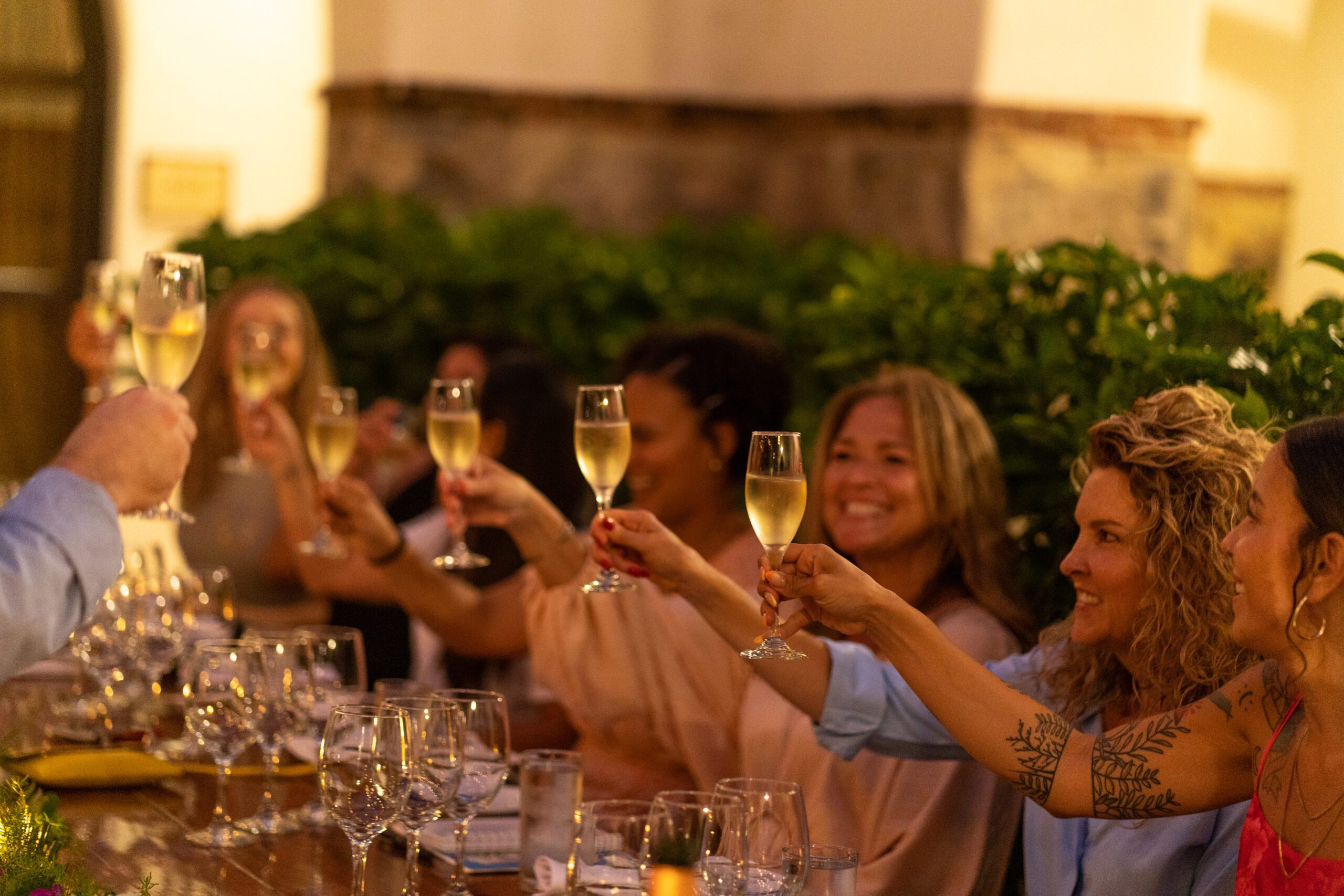 A group of people sit around a table outdoors at night, raising glasses of champagne in a toast, smiling and celebrating together at a festive Pop-Up Wine evening. The setting is warmly lit, creating the perfect ambiance for this special gathering.